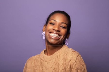 Téléchargez les photos : Charmante afro américaine en pull beige sourit happilye. Appréciation et confiance, concept de bonheur. Portrait sur fond de studio violet. - en image libre de droit
