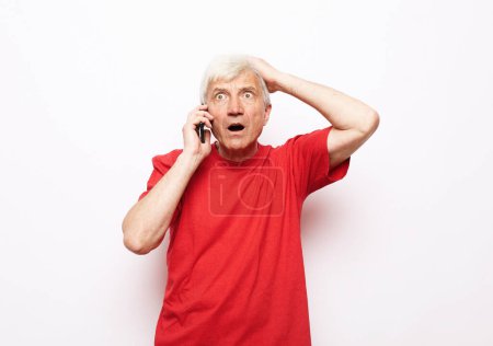 Téléchargez les photos : Style de vie moderne, tehnologie et concept de personnes âgées : Un homme âgé vêtu d'un T-shirt rouge tient un smartphone et entend de mauvaises nouvelles. Le pensionné est choqué. - en image libre de droit