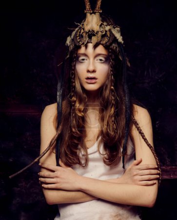Téléchargez les photos : Jeune femme avec un maquillage lumineux, des tresses et un crâne sur la tête se tient avec les bras croisés sur un fond noir - en image libre de droit