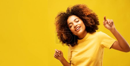 Foto de Estilo de vida, emoción y concepto de personas: retrato de mujer africana joven con gran pelo rizado afro sobre fondo amarillo bailando y con peinado volando en el aire - Imagen libre de derechos