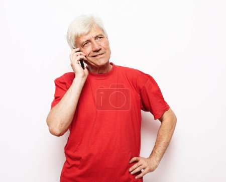 Foto de Estilo de vida moderno, tehnología y concepto de la gente mayor: Un anciano vestido con una camiseta roja sostiene un teléfono inteligente y escucha buenas noticias. Pensionista sonríe y se regocija de pie sobre un fondo blanco.. - Imagen libre de derechos