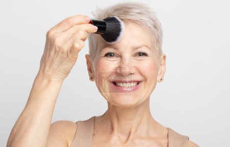 Photo for Beauty portrait of elderly woman applying blush with a makeup brush over grey background. Close up. - Royalty Free Image