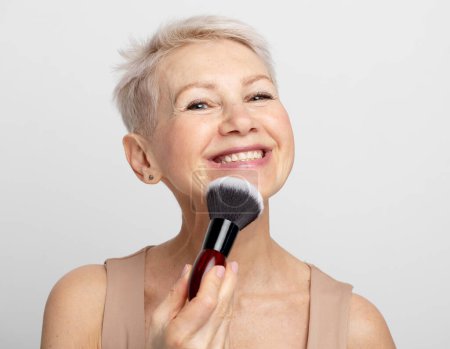 Photo for Beauty portrait of elderly woman applying blush with a makeup brush over grey background. Close up. - Royalty Free Image