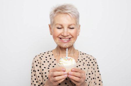 Téléchargez les photos : Portrait de femme âgée tenant cupcake d'anniversaire faisant vœu avec les yeux fermés et soufflant bougie célébrant anniversaire. Heureuse dame mature avec gâteau de fête - en image libre de droit
