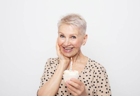 Foto de Alegre anciana con corte de pelo corto sostiene una magdalena con una vela en las manos, celebra un cumpleaños y ríe, sintiéndose feliz. - Imagen libre de derechos