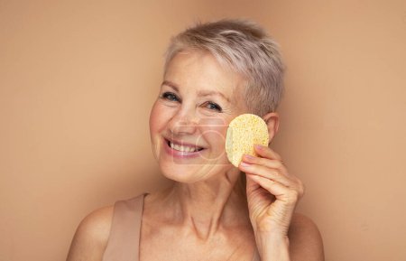 Téléchargez les photos : Photo de belle vieille femme utiliser éponge enlever le maquillage sur fond beige, gros plan - en image libre de droit
