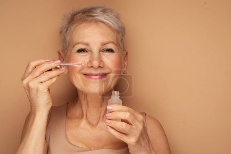 Foto de Happy senior lady aplica suero de aceite cosmético en la cara cuida la piel y sonríe ampliamente disfruta de tratamientos de belleza se queda siempre joven bellas poses sobre fondo beige - Imagen libre de derechos