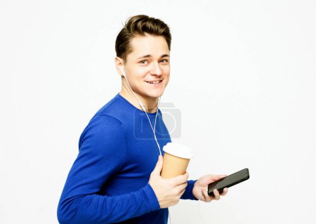 Photo for Smiling young man in blue sweater holds phone and cup of coffee to go, listening musing and feels happy. - Royalty Free Image