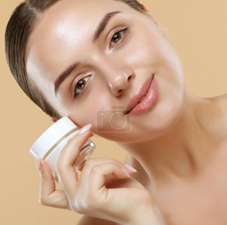 Photo for Beauty Youth Skin Care Concept - Beautiful Caucasian Woman Face Portrait holding and presenting cream jar product. Beautiful Spa model Girl with Perfect Fresh Clean Skin over beige background. - Royalty Free Image
