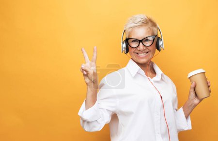Téléchargez les photos : Joyeux pensionné femme avec coupe de cheveux courte écoute de la musique et tient un verre de café, danse sur un fond jaune, se sent heureux. - en image libre de droit