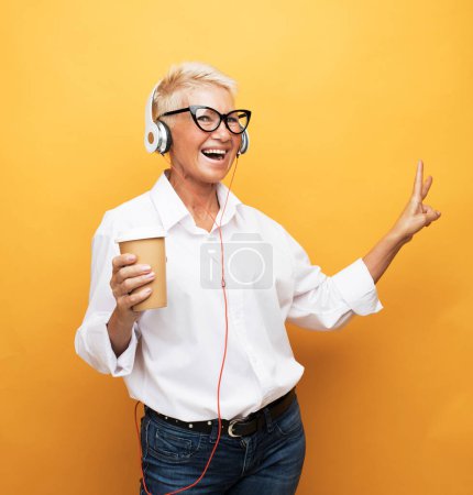 Téléchargez les photos : Portrait de femme âgée avec écouteurs écoutant de la musique et tenant une tasse de café sur fond jaune - en image libre de droit