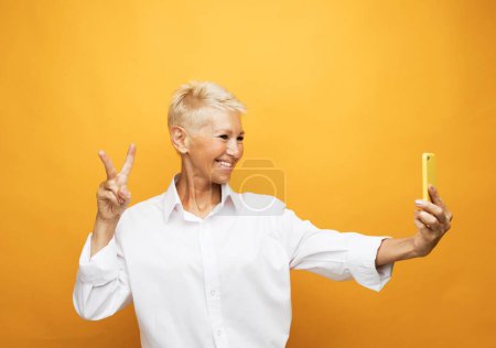 Photo for It's selfie time. Image of cheerful mature old woman standing isolated over yellow background wall talking by mobile phone. - Royalty Free Image
