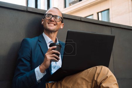 Foto de Joven con estilo en gafas trabaja con un ordenador portátil mientras está sentado en un banco en un entorno urbano. Empresario bebiendo café y charlando en internet - Imagen libre de derechos