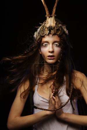 Photo for A young woman with bright make-up, braided hair and a skull on her head stands with her palms folded in prayer on a black background. Carnival party, halloween concept. - Royalty Free Image