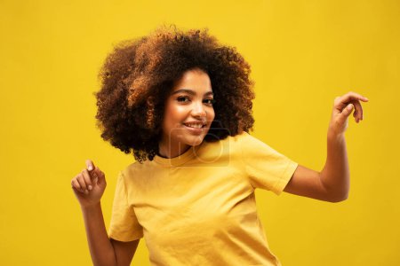 Photo for Positive dark skinned woman dances carefree raises arms moves with rhythm of favorite music dressed casually isolated over yellow background. Afro American female model makes triumph dance. - Royalty Free Image