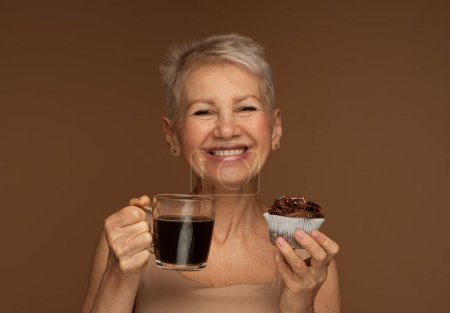 Photo pour Vieillesse, nourriture, boisson et concept de personnes - femme âgée souriante avec tasse de café et cupcake au chocolat sur fond brun. - image libre de droit