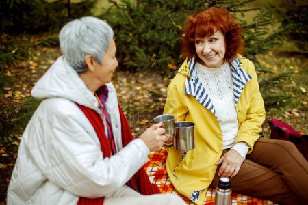 Téléchargez les photos : Two elderly women are sitting on a plaid blanket in the forest, drinking coffee, talking, having fun. Autumn time, happy old age. - en image libre de droit
