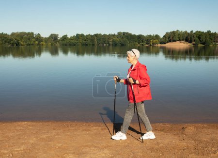 Foto de Deportiva mujer mayor entrena resistencia mientras nórdica caminar en la naturaleza, cerca del lago - Imagen libre de derechos