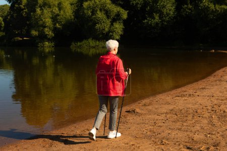 Téléchargez les photos : Grey-haired woman walking with tracking sticks on the beach near lake. Sport, healthy lifestyle and happy old age concept. - en image libre de droit
