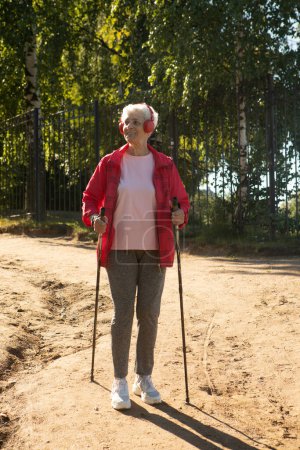 Photo for Summertime, active lifestyle, leisure and hobby concept. Outdoor shot of healthy energetic elderly female in red jacket and headphones walking on the beach sunny day using Nordic poles. - Royalty Free Image