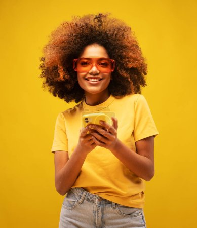 Photo for Portrait of lovely ethnic woman holds modern mobile phone, uses electronic device on surfing web, looks positively at camera, connected to wireless internet, wears yellow shirt and big sunglasses - Royalty Free Image