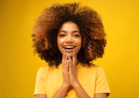 Téléchargez les photos : Jeune femme africaine surprise souriant à la caméra sur fond jaune. Jeune femme tient la main près de son visage et sourit largement. Concept de beauté. - en image libre de droit