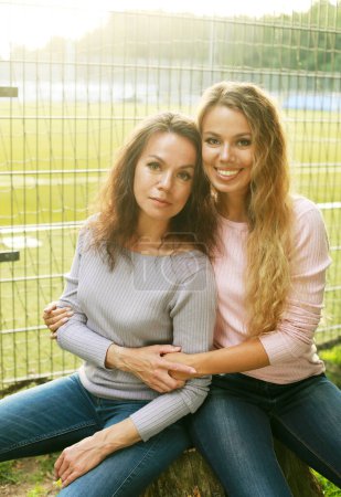 Téléchargez les photos : Two women are sitting on the grass in the park next to the football field. Beautiful young woman and her adult mother. Happy time. Two generations. - en image libre de droit