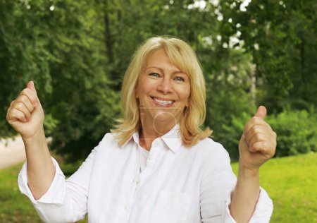 Téléchargez les photos : Charming blonde woman of sixty years in a summer park shows an ok sign, close-up portrait. The lady smiles and feels happy. - en image libre de droit