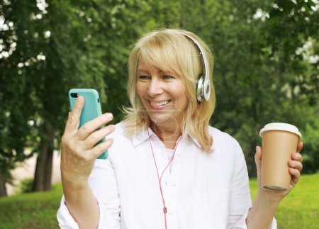 Photo for Beautiful elderly pensioner woman in headphones holding coffee to go and smartphone. Takes a selfie. Having fun in retirement on a summer afternoon. Golden age. - Royalty Free Image