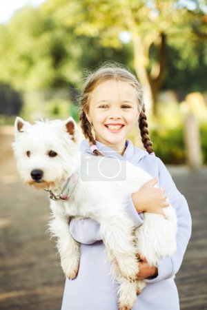 Téléchargez les photos : Animal friendship and happy childhood concept. A girl with pigtails holds a small white dog in her arms and smiles happily. - en image libre de droit