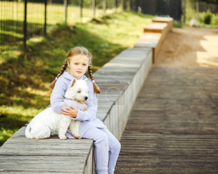 Téléchargez les photos : Animal friendship and happy childhood concept. The girl sits on a bench and hugs a small white dog. Summer day. - en image libre de droit