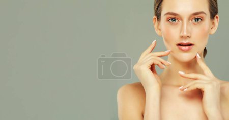 Foto de Encantadora joven con piel limpia y saludable y maquillaje natural. Retrato de cerca sobre fondo gris. La modelo femenina toca su cara. - Imagen libre de derechos