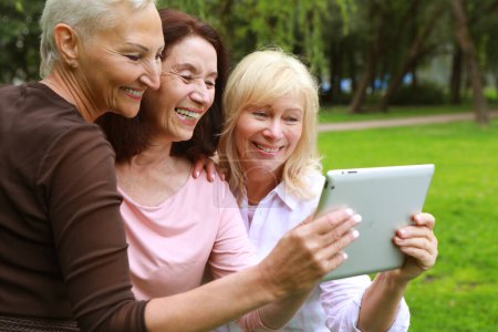 Foto de Tres ancianas sonríen y ven las noticias en la pantalla de la tableta en el parque en un día de verano. El concepto de amistad, feliz vejez y emociones. - Imagen libre de derechos