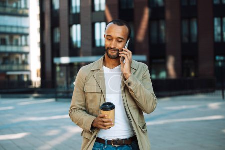 Photo for Young afro american man dressed casual holding a cup of coffee to go, smiling happy using smartphone at the city. Lifestyle and people concept. - Royalty Free Image