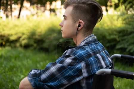 Téléchargez les photos : A young man in a wheelchair in the park listens to music, the guy is dressed in a plaid shirt, has piercings, an informal appearance. Side view, profile. - en image libre de droit