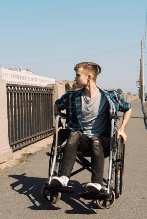Foto de Un joven discapacitado cabalga en una silla de ruedas a través de un puente, el joven hombre rubio vestido con una camisa a cuadros y jeans. - Imagen libre de derechos