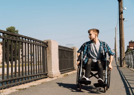 Un joven discapacitado cabalga en una silla de ruedas a través de un puente, el joven hombre rubio vestido con una camisa a cuadros y jeans.