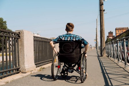 Foto de Un joven discapacitado cabalga en una silla de ruedas a través de un puente, el joven hombre vestido con una camisa a cuadros y vaqueros. Vista trasera. - Imagen libre de derechos