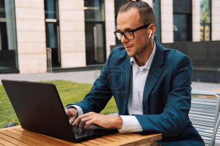 Foto de Joven hombre hipster con anteojos sentados en la mesa y escribiendo en el teclado del ordenador portátil mientras trabaja en la cafetería al aire libre - Imagen libre de derechos