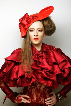 Foto de Concepto de fiesta, moda y gente: Joven mujer hermosa en un traje de encaje rojo y sombrero. Una elegante rubia escarlata, símbolo de pasión y energía, posa sobre un fondo gris claro. - Imagen libre de derechos