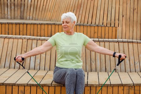 Foto de Old woman with short hair wear green t-shirt doing exercises outdoor , sitting on wooden bench using resistance rubber bands. Healthy lifestyle, active retired life and sporty time. - Imagen libre de derechos