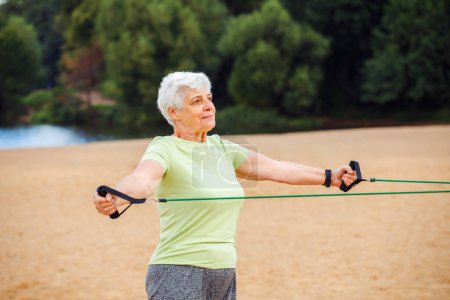 Photo for Old woman with short hair wear sportswear doing exercises outdoor on the beach in the morning using resistance rubber bands. Healthy lifestyle, active retired life and sporty time. - Royalty Free Image
