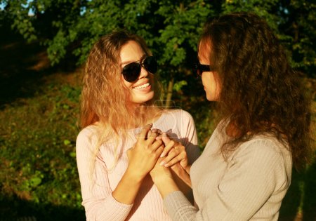 Senior Mom and her adult daughter hold hands in a summer park, happy women wearing sunglasses