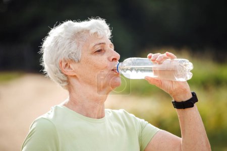 Senior woman with short grey hair drinking water after exercising, summer time, portrait in the park