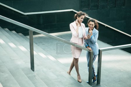 Photo for Business, people and tehnology concept: Two young successful business women smile and watch news on a smartphone, use social networks. They stand on the street, on the stairs on a summer day. - Royalty Free Image