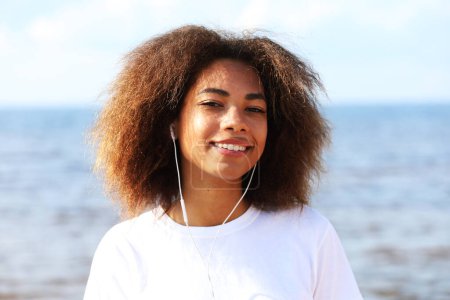 Foto de Encantadora joven afroamericana increíble escuchando música en auriculares en su teléfono móvil. Vestido casual. - Imagen libre de derechos