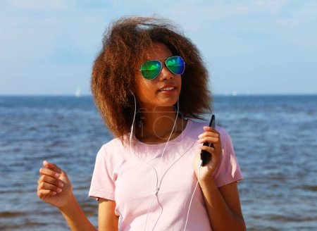 Foto de Mujer afroamericana joven con camiseta rosa en gafas de sol escuchando música en auriculares en su teléfono móvil cerca del mar. Concepto de estilo de vida. - Imagen libre de derechos