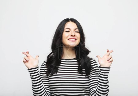 Photo for Lifestyle, emotion and people concept: Joyful hopeful and charismatic young brunette woman smiling cross fingers for dream come true over grey background - Royalty Free Image