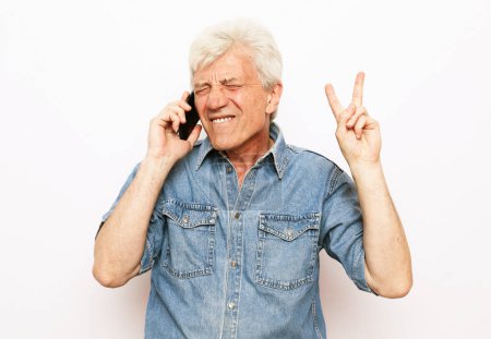 Téléchargez les photos : Style de vie moderne, tehnologie et concept de personnes âgées : Un homme âgé vêtu d'une chemise en denim tient un smartphone et entend de bonnes nouvelles. Pensionné sourit et se réjouit en se tenant debout sur un fond blanc.. - en image libre de droit