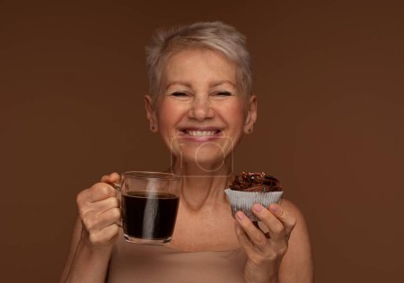 Téléchargez les photos : Vieillesse, nourriture, boisson et concept de personnes - femme âgée souriante avec tasse de café et cupcake au chocolat sur fond brun. - en image libre de droit